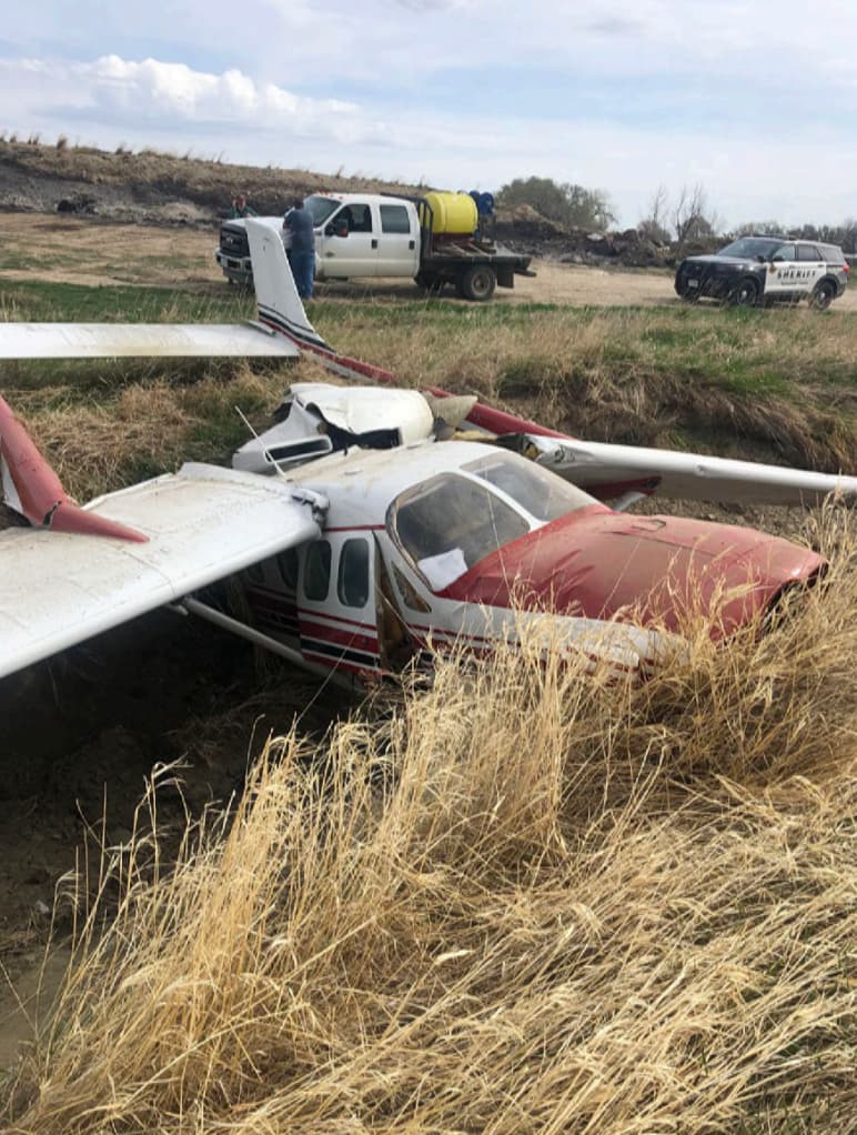 Close-up of the Cessna P337H N62PC cockpit and wing resting in a ditch, with oil stains on fuselage. Emergency responders and sheriff vehicle in background.